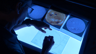 A person drawing on a piece of paper in the darkness. Three other pieces of paper show the sun.