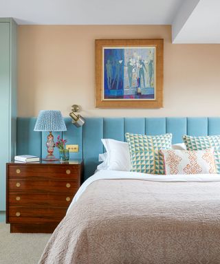 Pink and blue bedroom with colorful cushions, upholstered headboard, artwork, table lamp on wooden bedside table