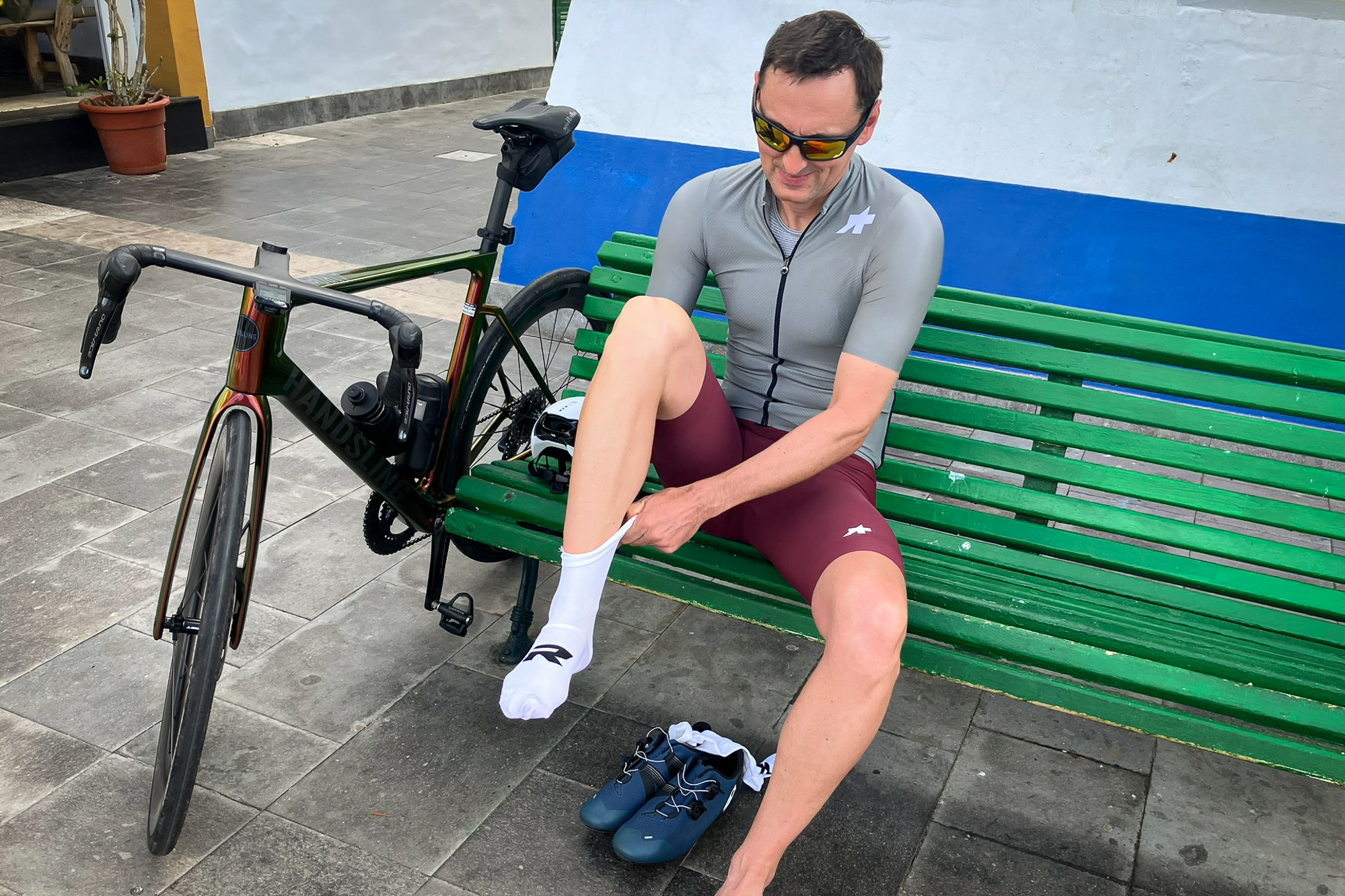 Man wearing a green cycling jersey and red shorts sittng on a green bench putting on some white socks.