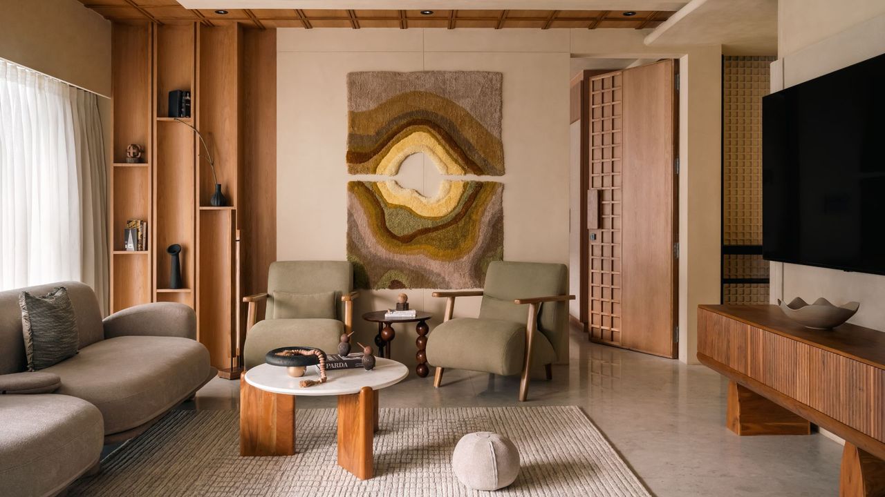 An earthy living room with beige walls, wood shelves, a beige rug, a round pillow, a coffee table with wood legs, a bobbin-style side table, a pair of gray armchairs, a gray sofa, a wood panelled sideboard by a TV, and a textured wall hanging