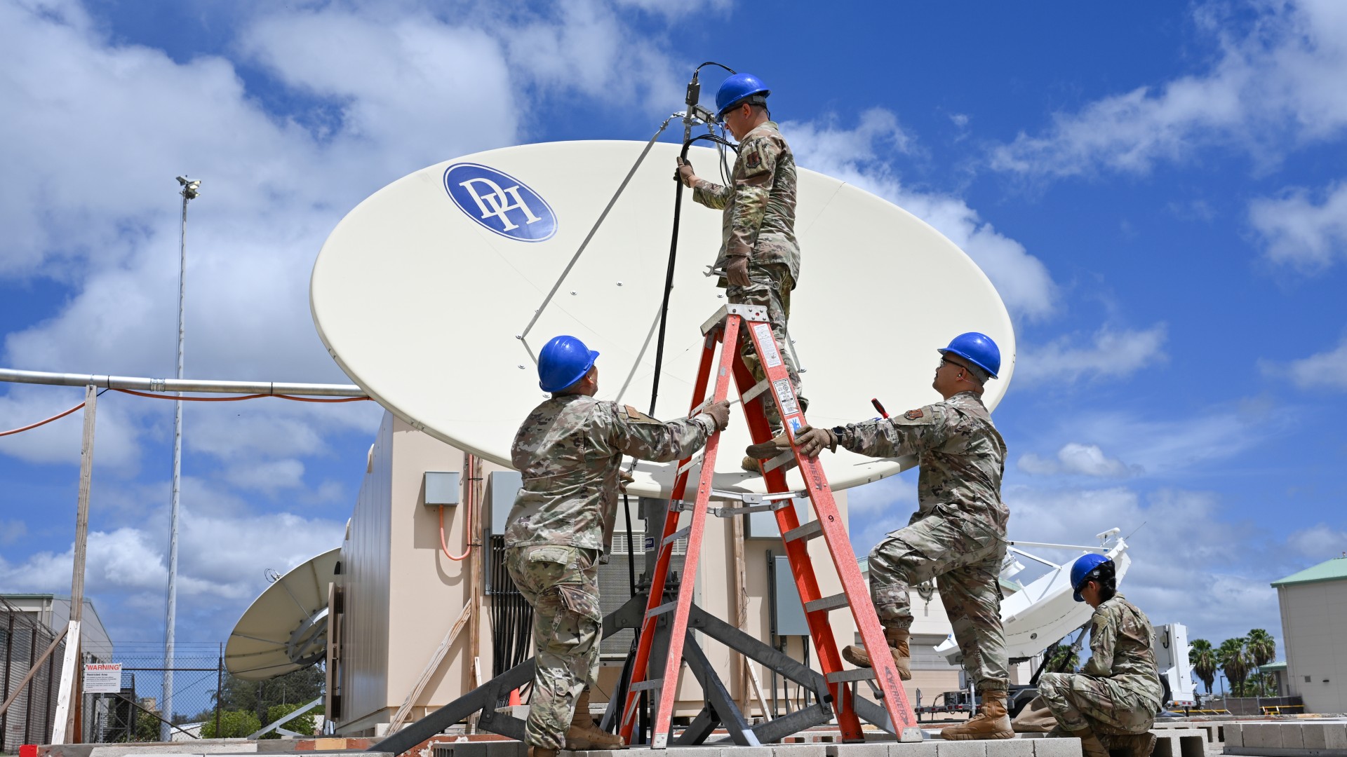 three people in camouflage uniforms stand beside a large satellite dish under a blue sky