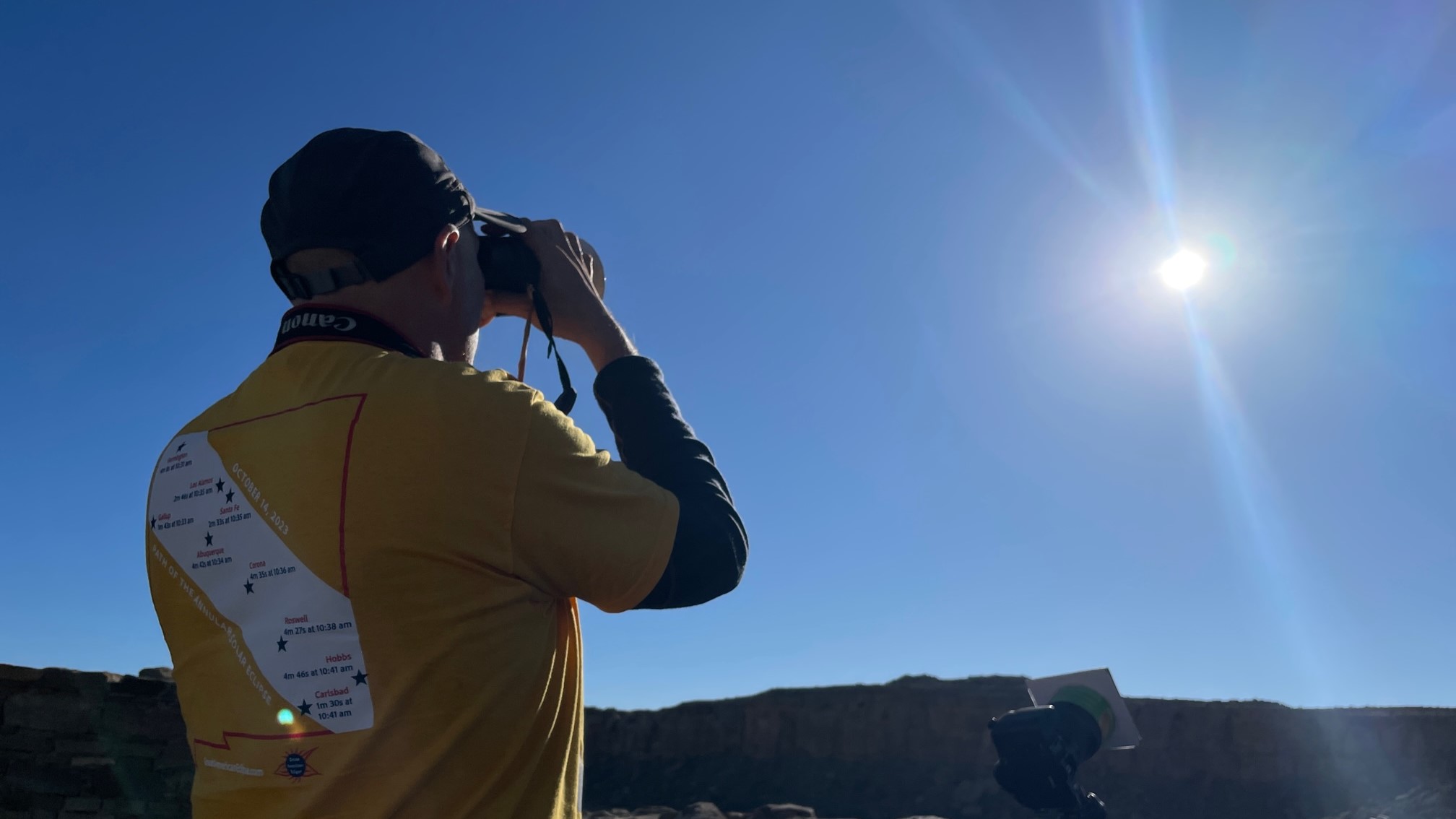A man wearing a yellow t shirt holds a pair of binoculars and looks at the sun.