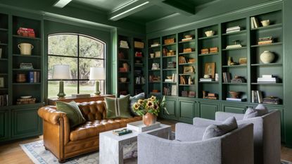 Green living room with built-in bookshelves filled with books and decor, a leather Chesterfield couch, and two grey armchairs.