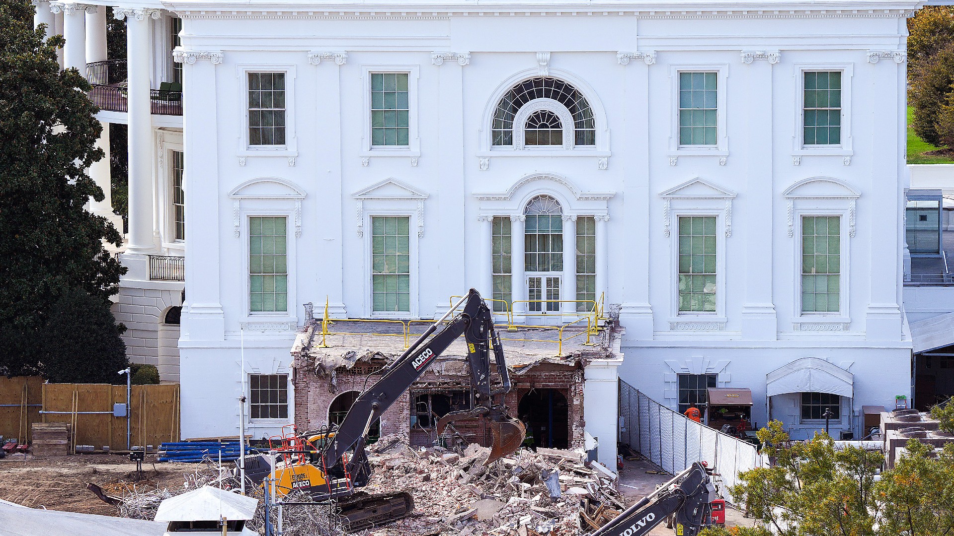 an excavator picks up rubble beside a white building
