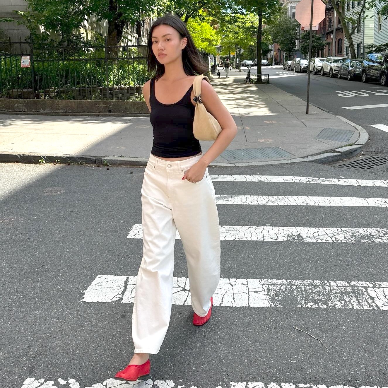 style influencer Mimi Nguyen walking across a street in New York City wearing a black tank top, tan suede bag, white barrel jeans, and red mesh ballet flats