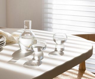 A clear carafe and two drinking glasses filled with water on a neutral colored dining table in a natural room. Light filters through a horizontal blind.