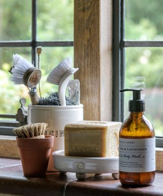 Close up of soap, jar of brushes and hand wash on a windowsill in front of a leaded window.