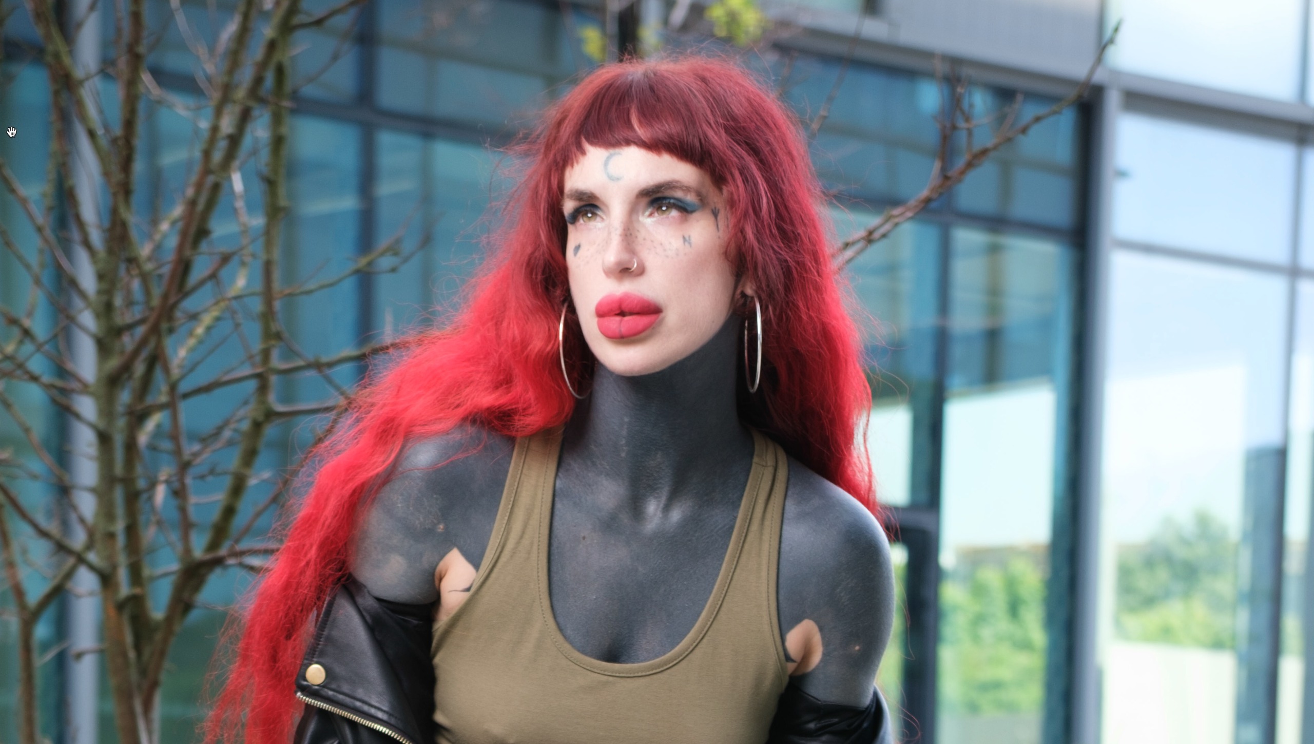 Red-haired person with dark body paint sitting on a wooden bench in black leather pants and boots outside a modern glass building.