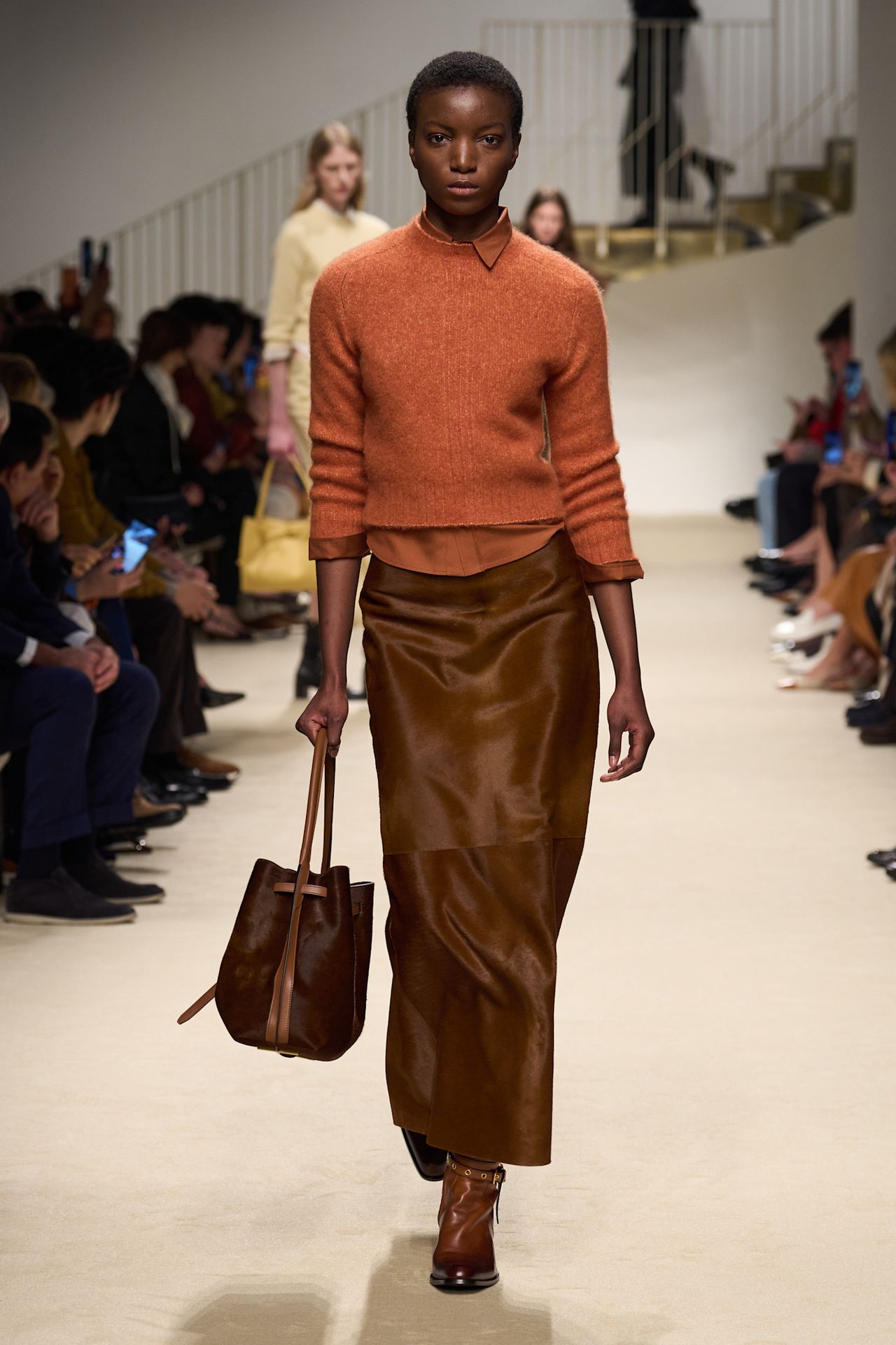 A Tod's spring/summer 2026 runway model wearing a rust orange shirt and sweater styled with a maxi-length brown pony hair pencil skirt and boots.