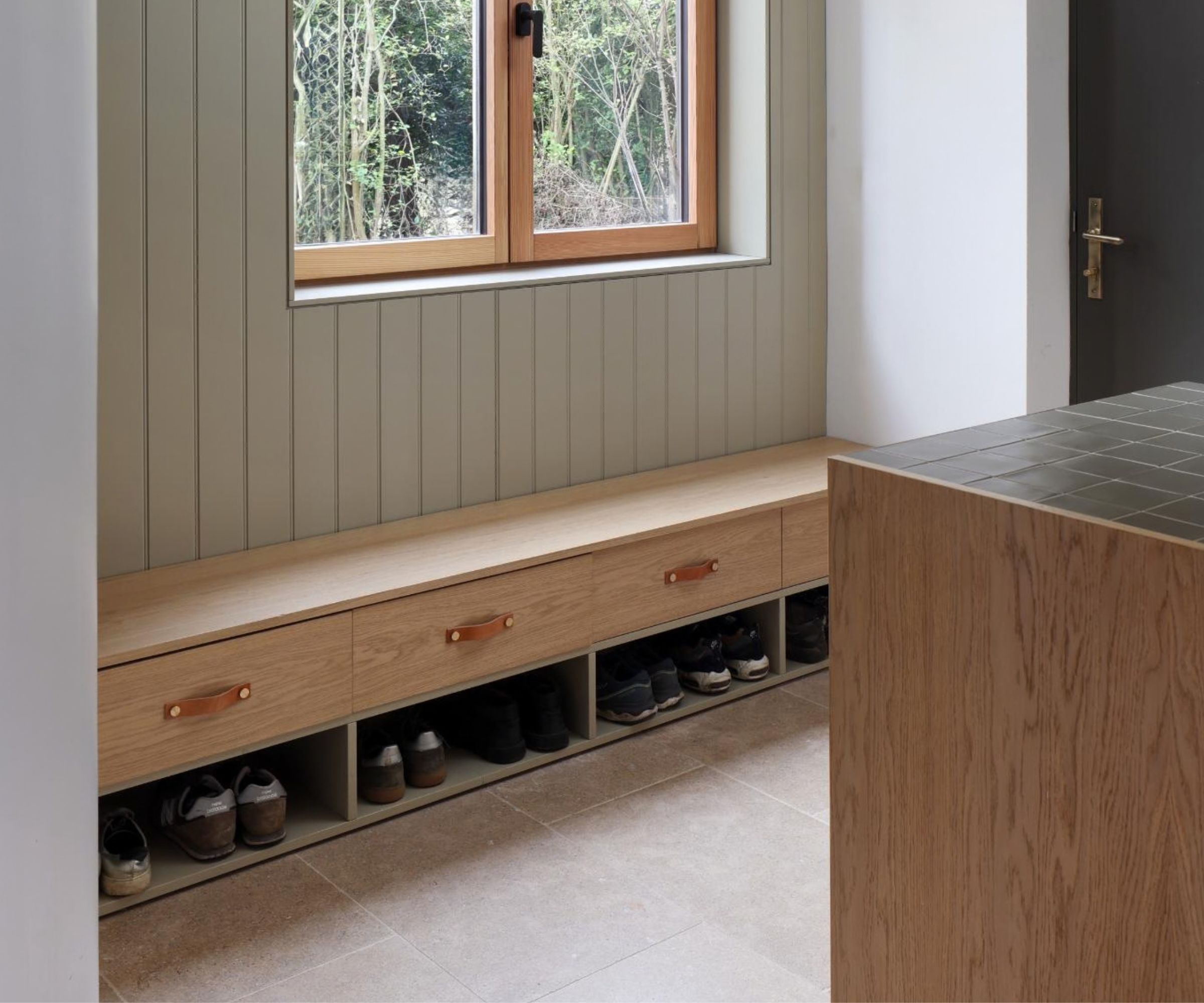 A built-in oak window seat with open cubby shoe storage below and three drawers with tan leather pull handles, set against sage green tongue-and-groove wall panelling in a country-style hallway with stone floor tiles.