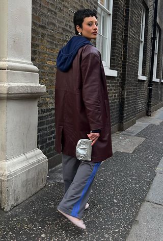 A woman walking down the sidewalk wearing a navy hooded sweatshirt under a dark red leather jacket with gray track pants, pink ballet flats, and a silver clutch bag.