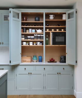 A light blue cabinet with open doors showcasing light wooden shelves with mugs, teapots, a microwave, and toaster within it, with cabinet drawers and cupboards underneath it