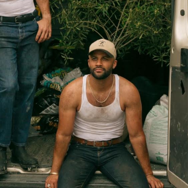 a headshot of co-founder of the plant daddies dane jordan, sitting on the edge of the back of a van filled with plants, wearing jeans, a white vest top covered in mud, a gold necklace and a beige cap staring off camera