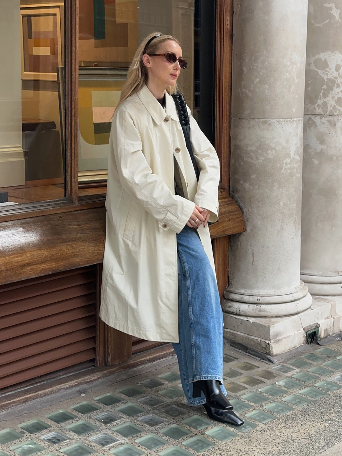 British style influencer Chloe Butler poses on a London sidewalk wearing a Carolyn Bessette-Kennedy-inspired outfit with a black headband, oval tort sunglasses, a light colored trench coat, bootcut jeans, and black square-toe boots