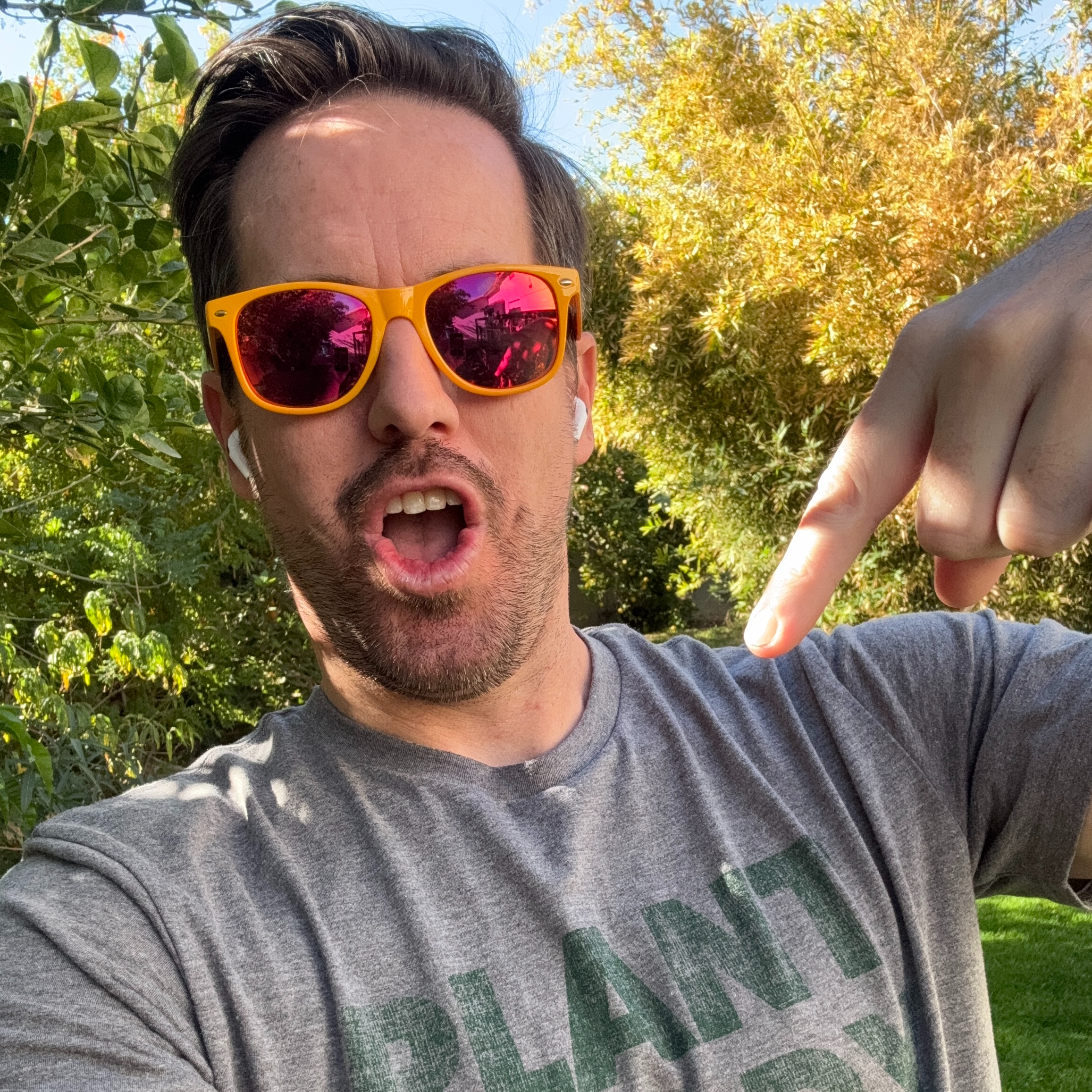 a headshot of plant expert Michael Overstreet: a man with dark hair in orange sunglasses standing outside surrounded by trees pointing at his plant-related shirt