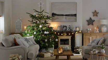 Pale grey living room, large decorated Christmas tree, grey sofa and armchair, festive cushions, wood burning stove, wooden reusable advent calendar. Pub Orig