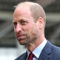 Prince William wearing a blue suit jacket, white shirt and red tie standing outside in the rain looking amused