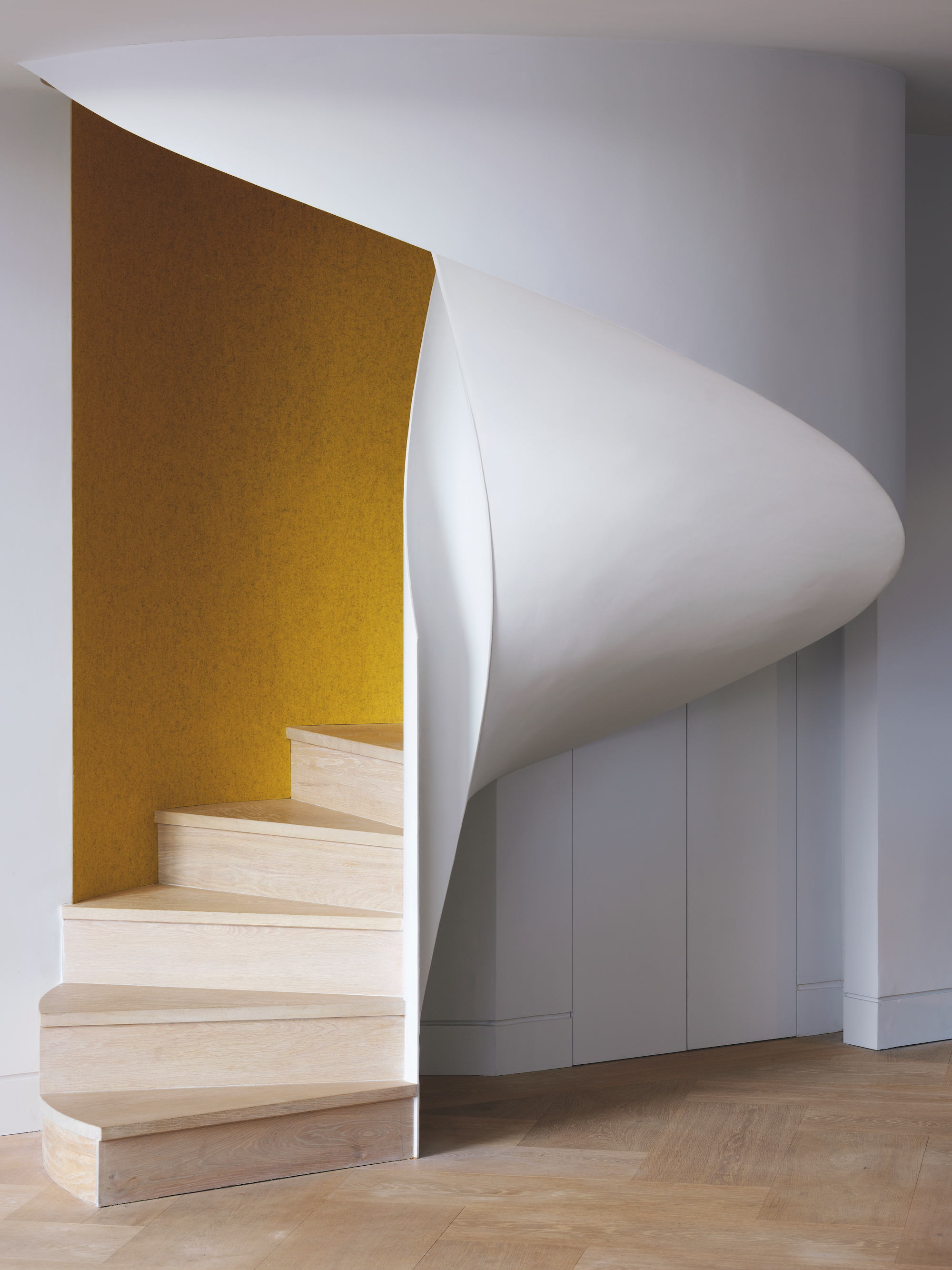 A white, plastered spiral staircase with a mustard yellow wall inside, and light toned wood stairs.