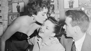 Screen star lovely Elizabeth Taylor bends low to kiss Judy Garland and wish her luck during a visit backstage at the Palace. Looking on at the buss business is actor Montgomery Clift, Miss Taylor's escort to the theater, where Judy is reviving the lost art of Vaudeville.