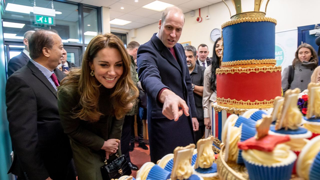 Prince William pointing at a blue cupcake and Princess Kate leaning over to look at it and smiling