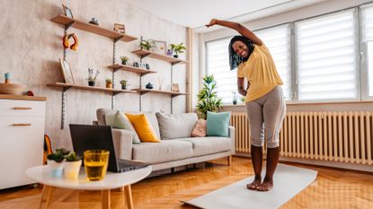 woman doing a side bend in a living room home setting on an exercise mat in front of a laptop on a side table, facing the camera and smiling