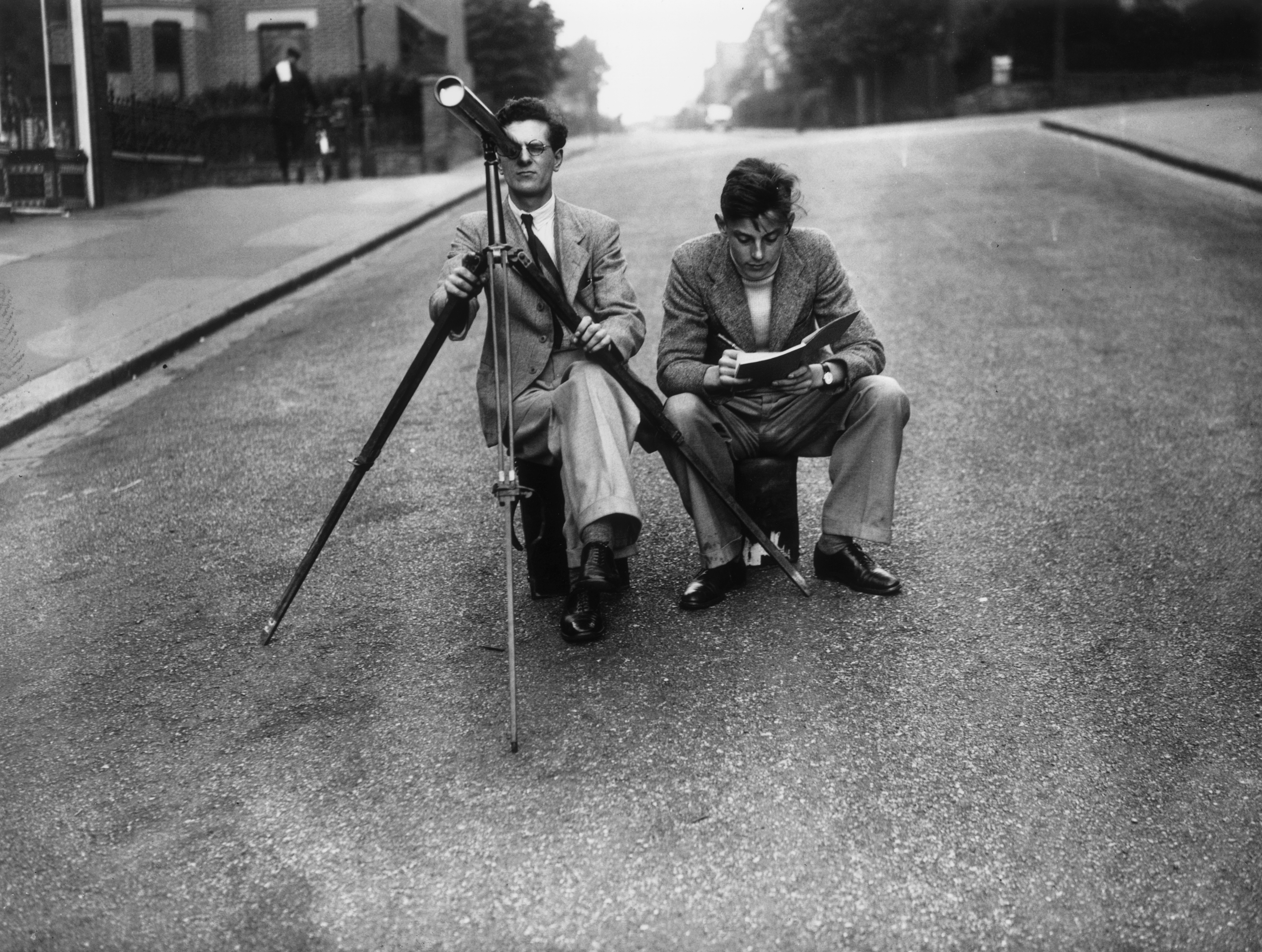 two men sit in the middle of the road, one man is looking through a telescope and the other is looking at a leaflet.