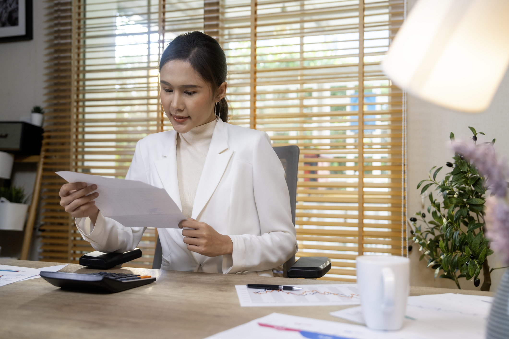 Woman looks at payslip on a piece of paper as she sits at a desk which has a calculator on it.