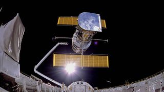 fisheye view of space shuttle payload bay with a robotic arm at center. in foreground is hubble space telescope with two solar panels floating into space