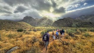 Hikers walking towards moody clouds with sunlight piercing through above a rugged landscape.