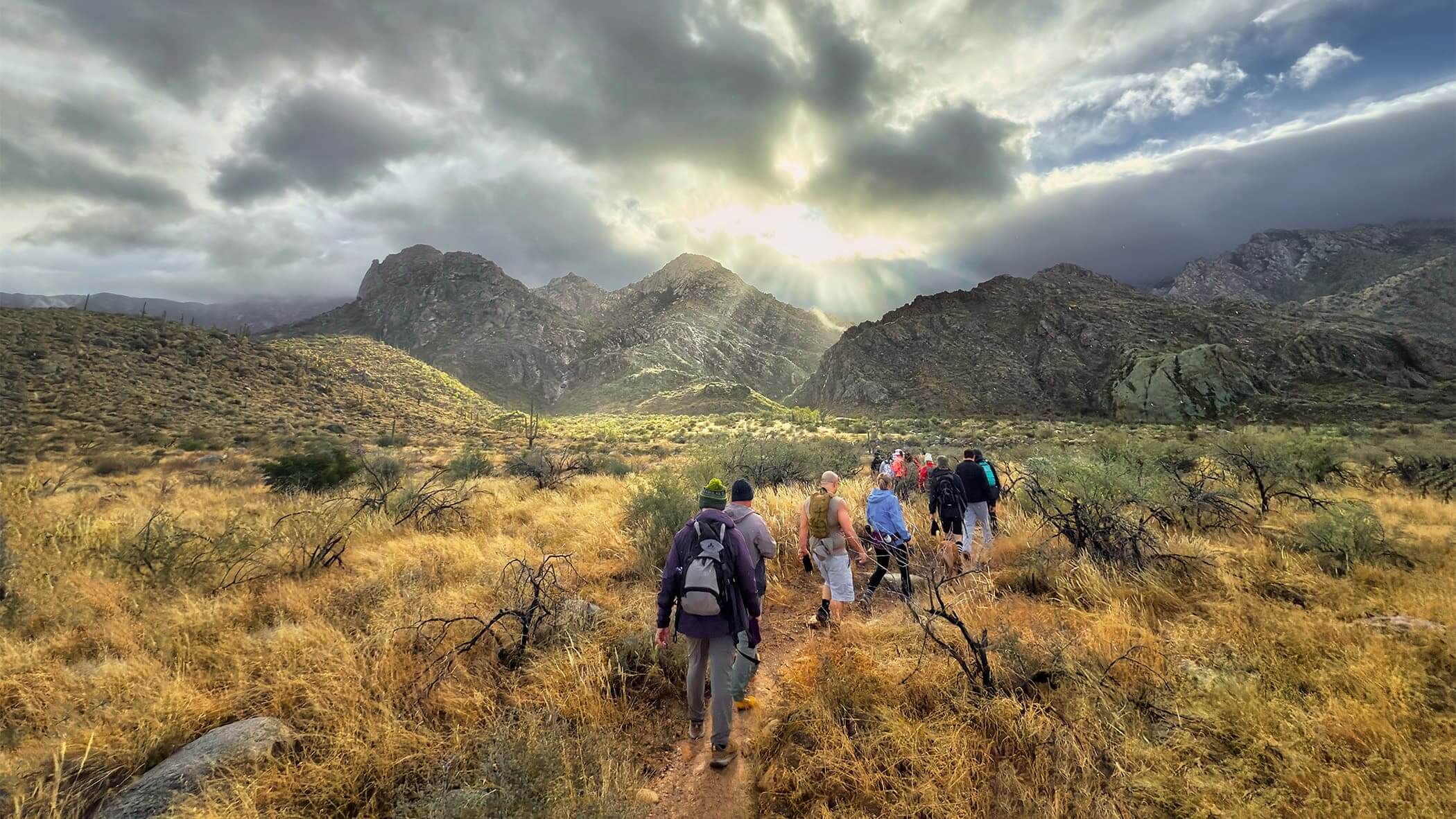 Hikers walking towards moody clouds with sunlight piercing through above a rugged landscape.