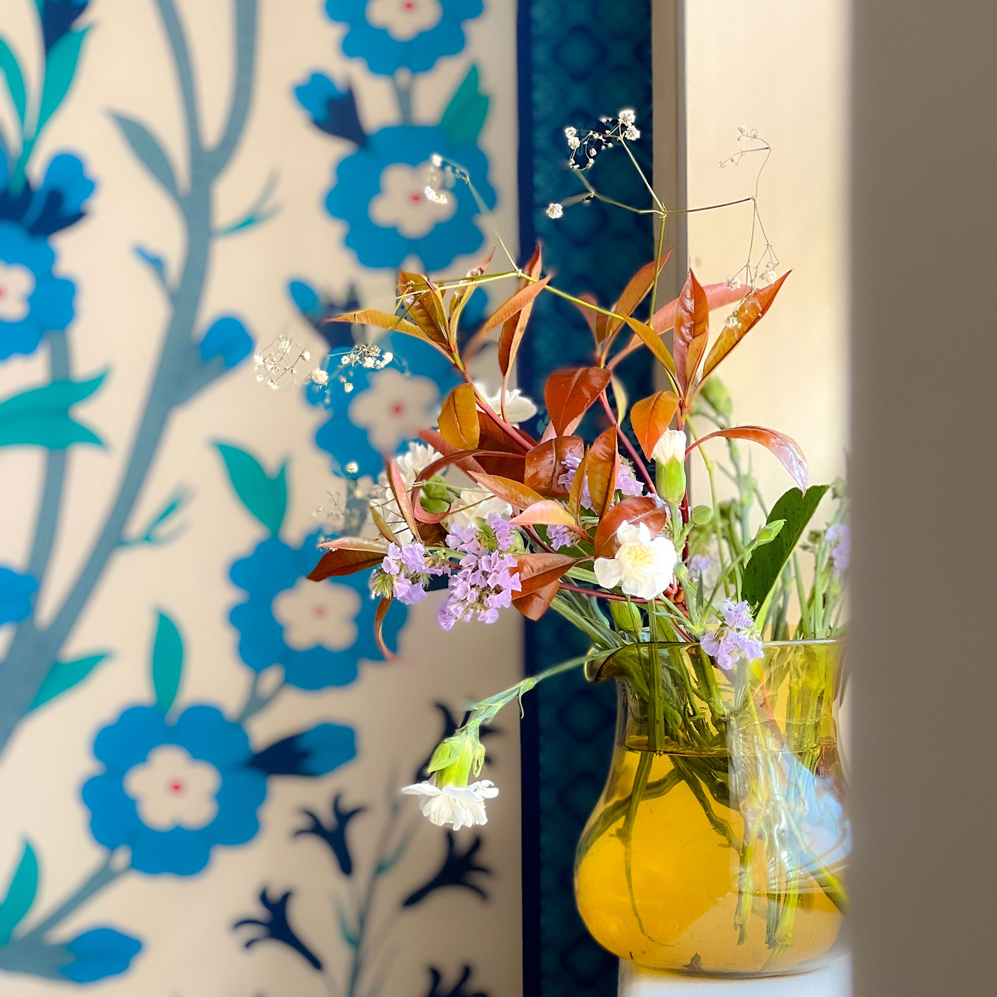 Yellow glass vase filled with fresh flowers in front of blue floral patterned backdrop