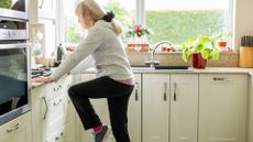 woman standing in the kitchen sideways to the camera raising her knee and holding onto the counter. she's wearing a cream hoodie and black trousers.
