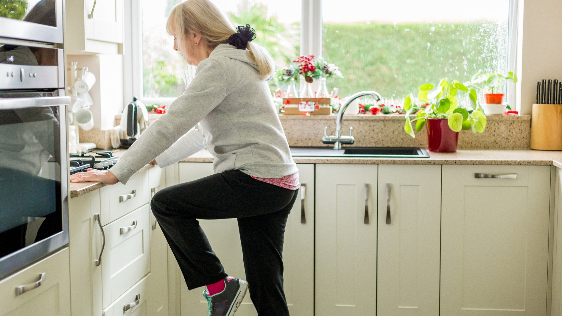 woman standing in the kitchen sideways to the camera raising her knee and holding onto the counter. she's wearing a cream hoodie and black trousers. 