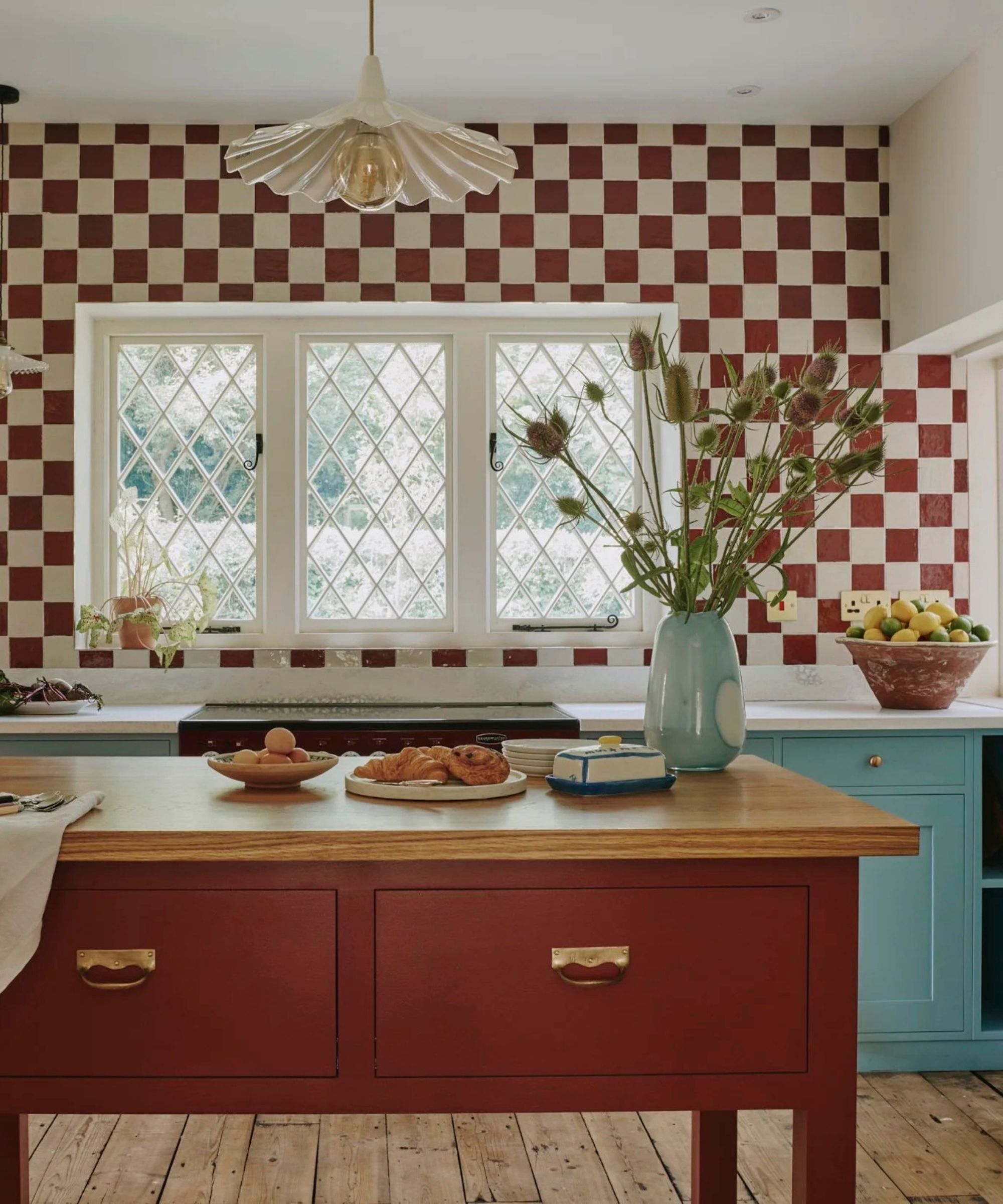 a kitchen with a checkerboard tile backsplash, lower blue painted cabinets, an island with a wooden top surface and red painted drawers, a vase of flowers on the island, and a pendant light hanging from the ceiling