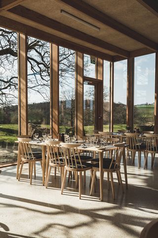 A sun-lit, wood and glass pavilion features Scandinavian interiors including linear wooden tables and matching chairs and floor-to-ceiling windows.