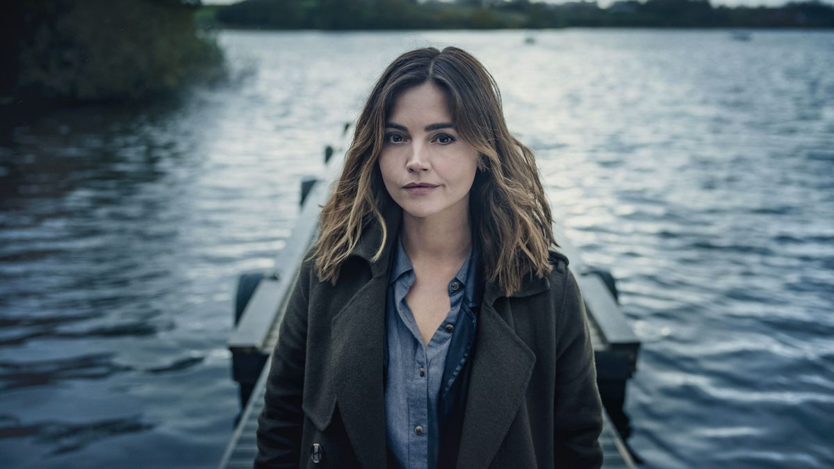 Jenna Coleman in a dark jacket as Ember Manning stands on a jetty with water in the background in The Jetty