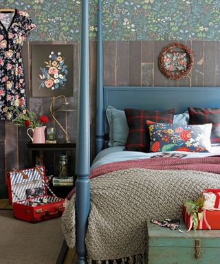 a bedroom with a blue four poster bed, wooly blankets and tartan cushions on top, a red vintage suitcase on the floor and a black floral dress hanging up on a wall that has a green floral wallpaper on top and on the bottom wallpaper with black and gold lined panels