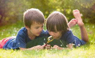 Two boys lying on grass explore nature with a magnifying glass.