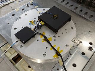 a light grey circular plate sits on a rectangular steel plate with various bolt holes punched in both. on the top plate, two black boxes of different sizes are connected to wires that rail out of frame.