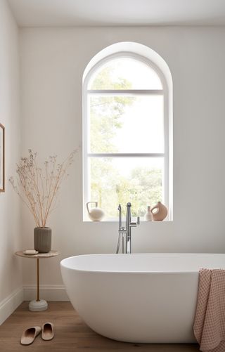 A white bathroom with a white curved bath, wooden floors and a circular table with dried flowers