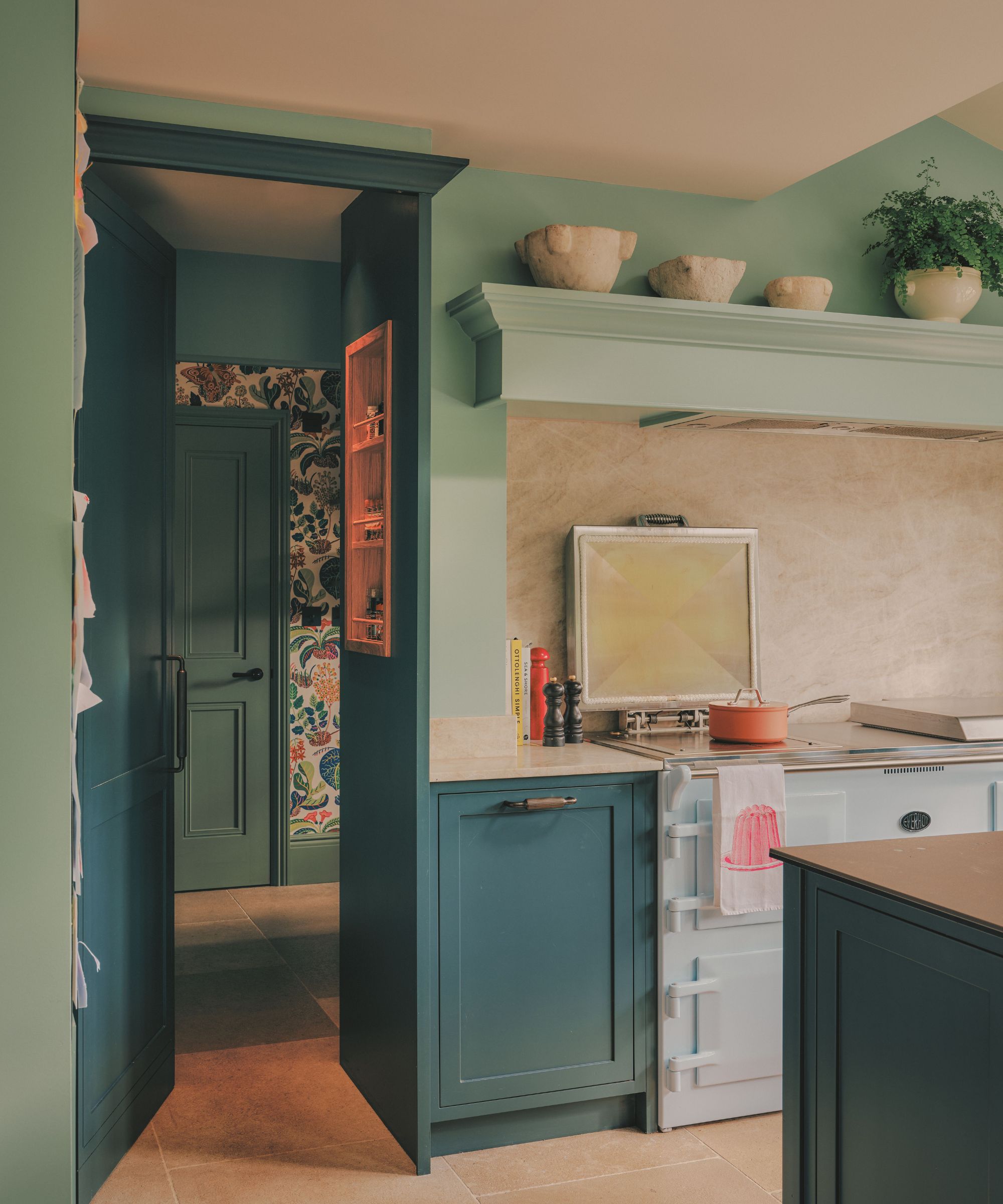 Sage green kitchen corner featuring teal blue cabinetry, an off-white range cooker, a stone splashback, and a glimpse into a room with floral wallpaper.
