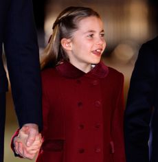 Princess Charlotte wearing a burgundy coat and smiling holding Prince William's hand
