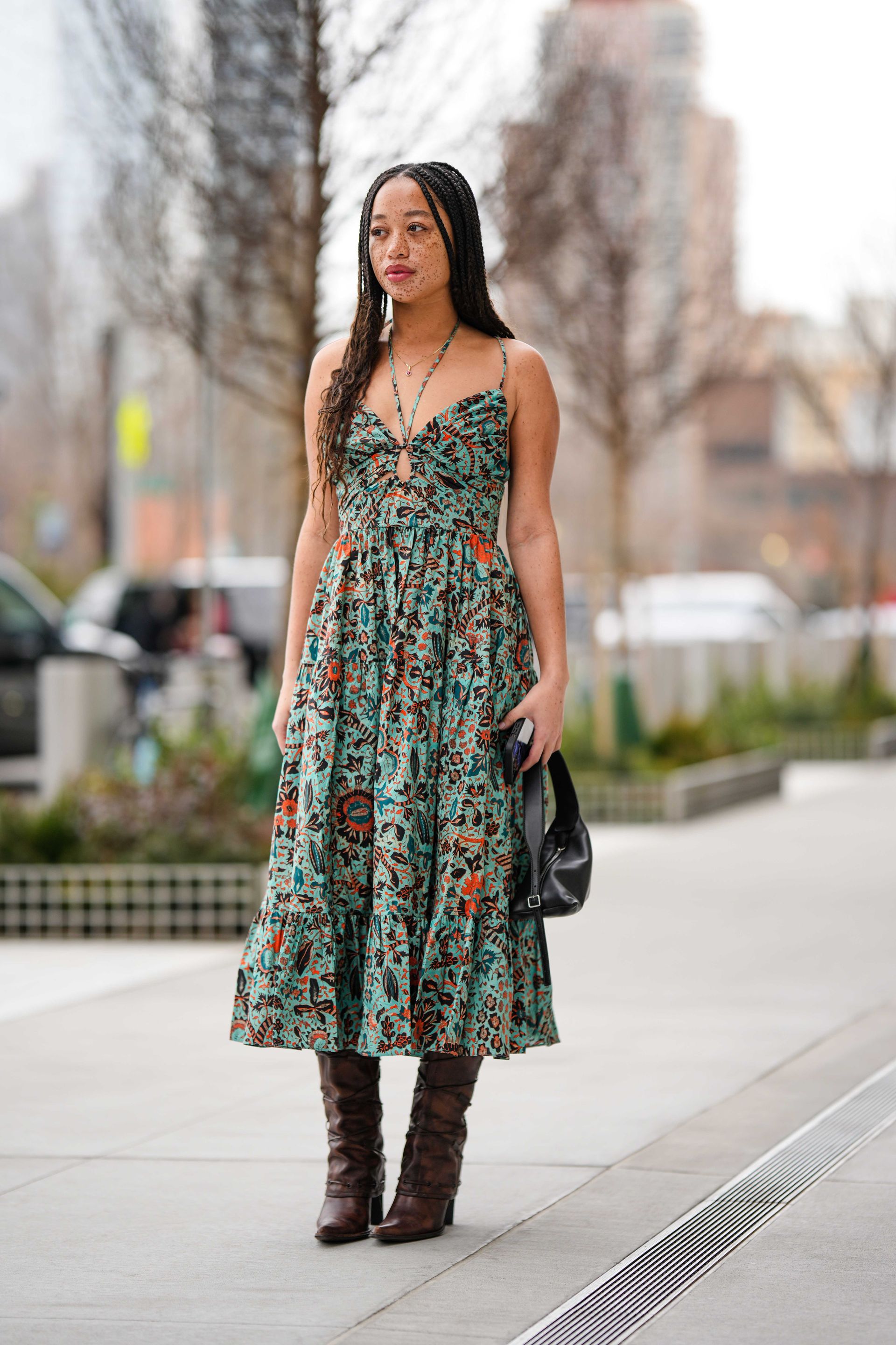A guest wears a black / pale blue / orange print pattern laces halter neck / tank-top / cut-out chest long dress, a black shiny leather micro handbag, dark brown shiny leather block heels / laces details / knees boots , outside Ulla Johnson, during New York Fashion Week, on February 12, 2023 in New York City