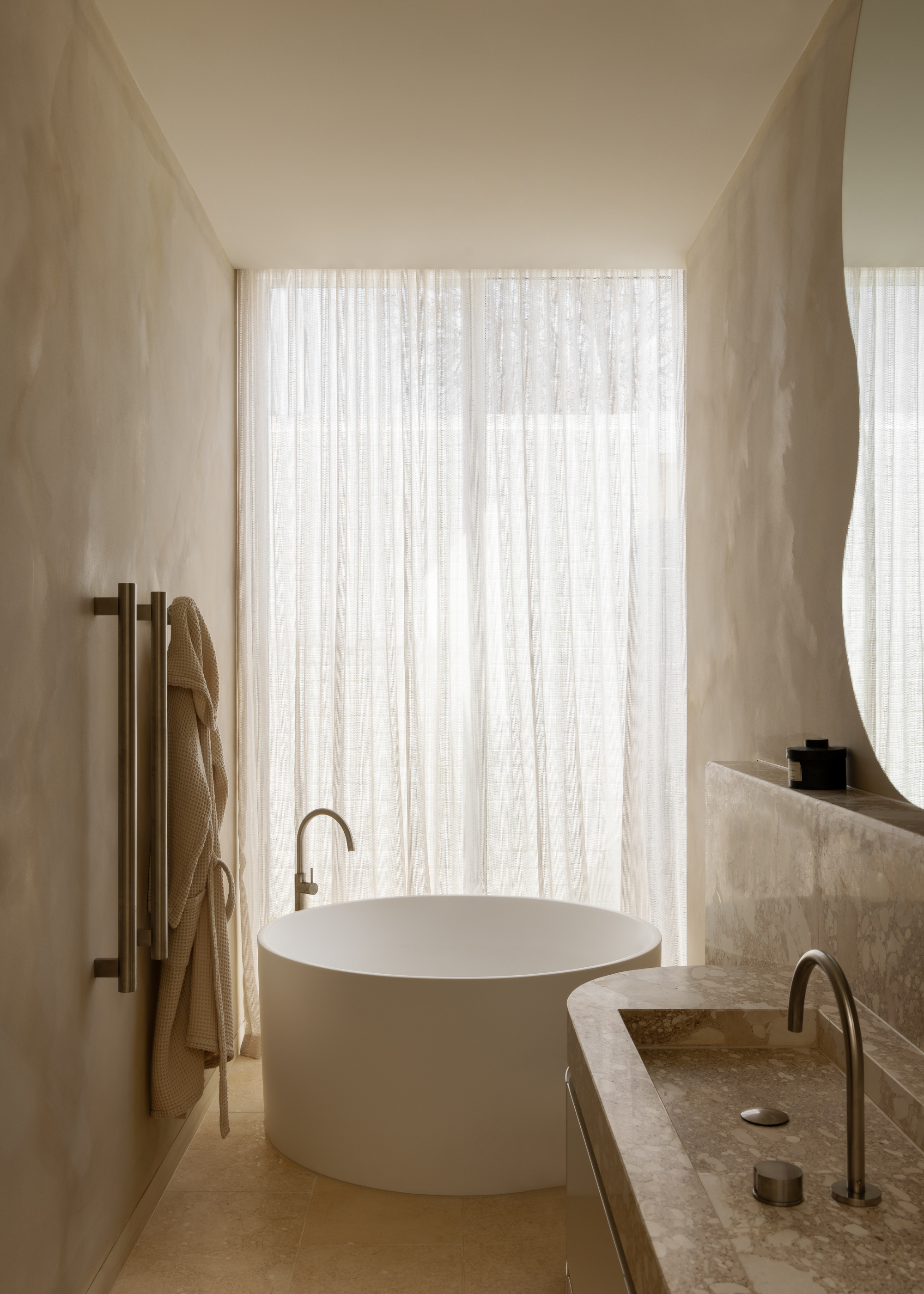 A beautiful bathroom with a circular tub, sheer curtains by a floor-length window, a hanging robe and a marble sink