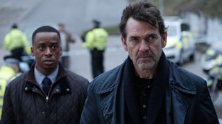 Elliot Edusah as DC Mike Dilliston and Dougray Scott and DI Lou Stanley in dark coats stand with uninformed police in the background in The Crow Girl.