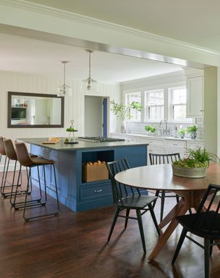 blue kitchen island with bar stools and oval wood dining table and black spindleback dining chairs and white walls, tiles and cabinets and a dark wood floor
