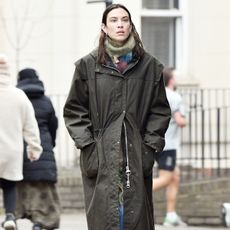 Alexa chung wears a green long coat, jeans, patent leather boots, and a plaid scarf.