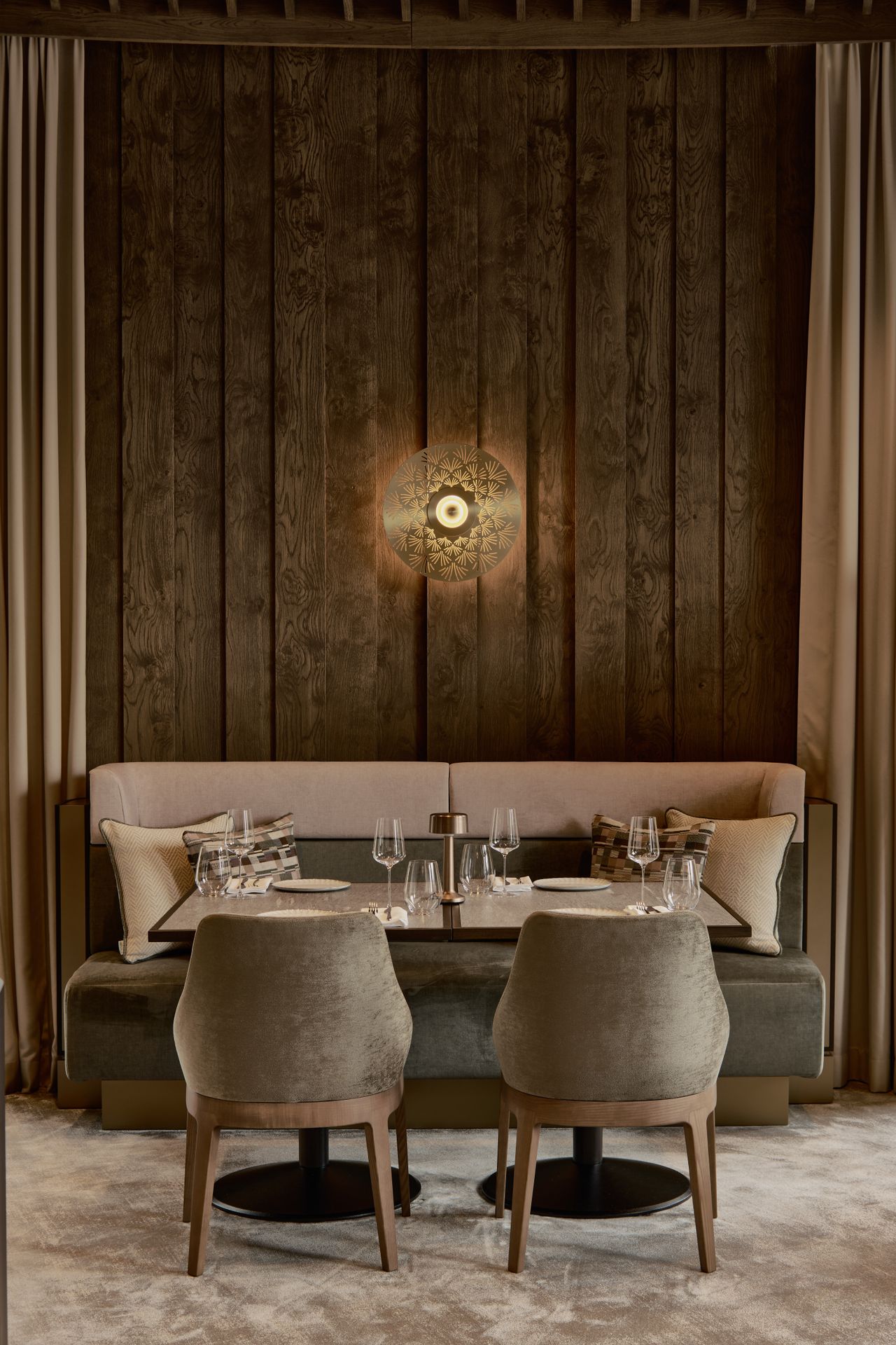 An earthily tinted restaurant dining room features plush furnishings, including upholstered velvety chairs in muted olive green, a brown, cream, and dark green banquette, and wood-paneled walls, lit up by a spherical wall light.