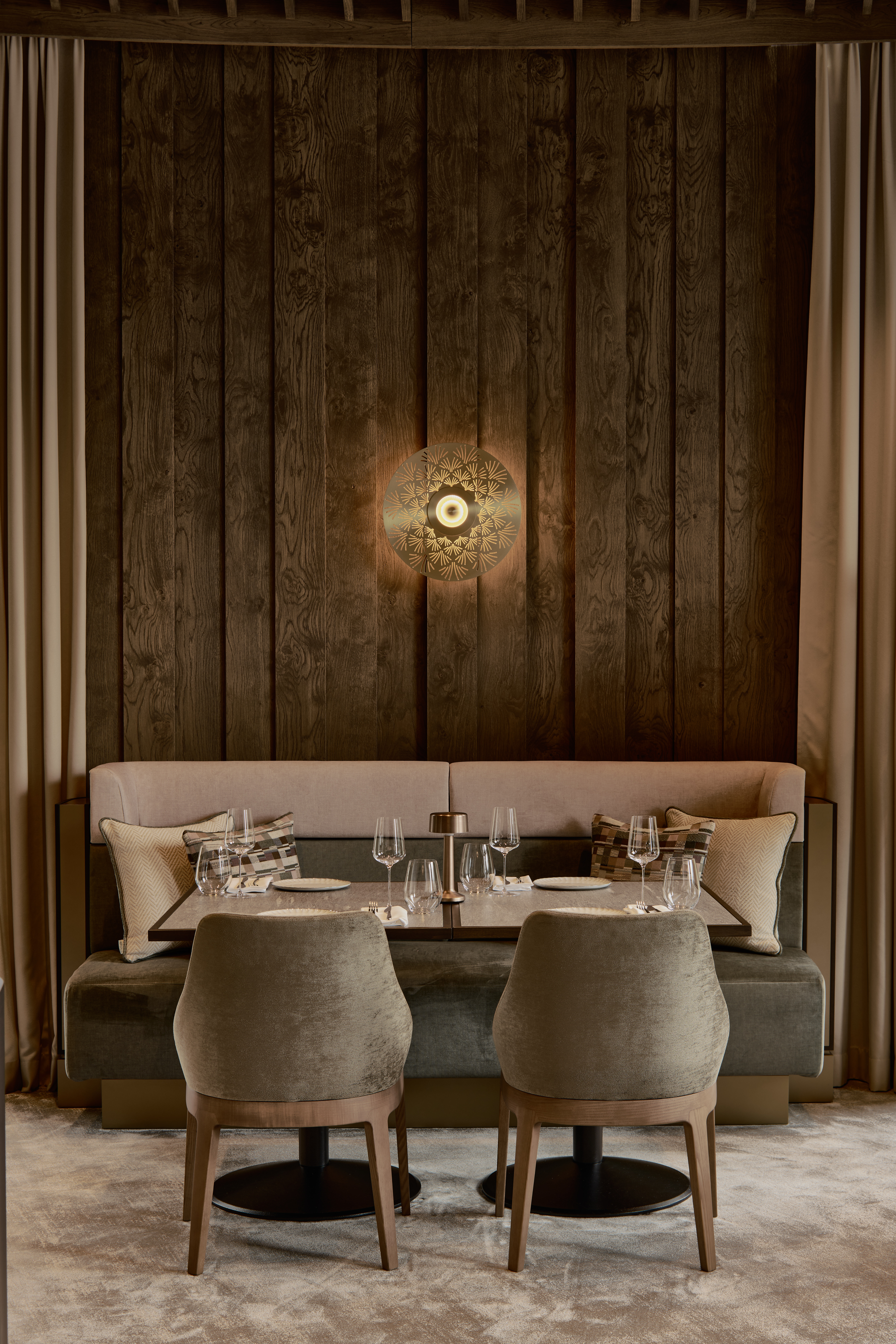 An earthily tinted restaurant dining room features plush furnishings, including upholstered velvety chairs in muted olive green, a brown, cream, and dark green banquette, and wood-paneled walls, lit up by a spherical wall light.