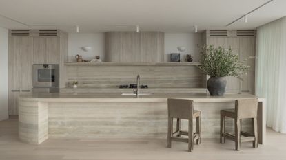 A beautiful kitchen with a long stone island, matching backsplash and exhaust, brown kitchen stools, and an earthen vase with stems
